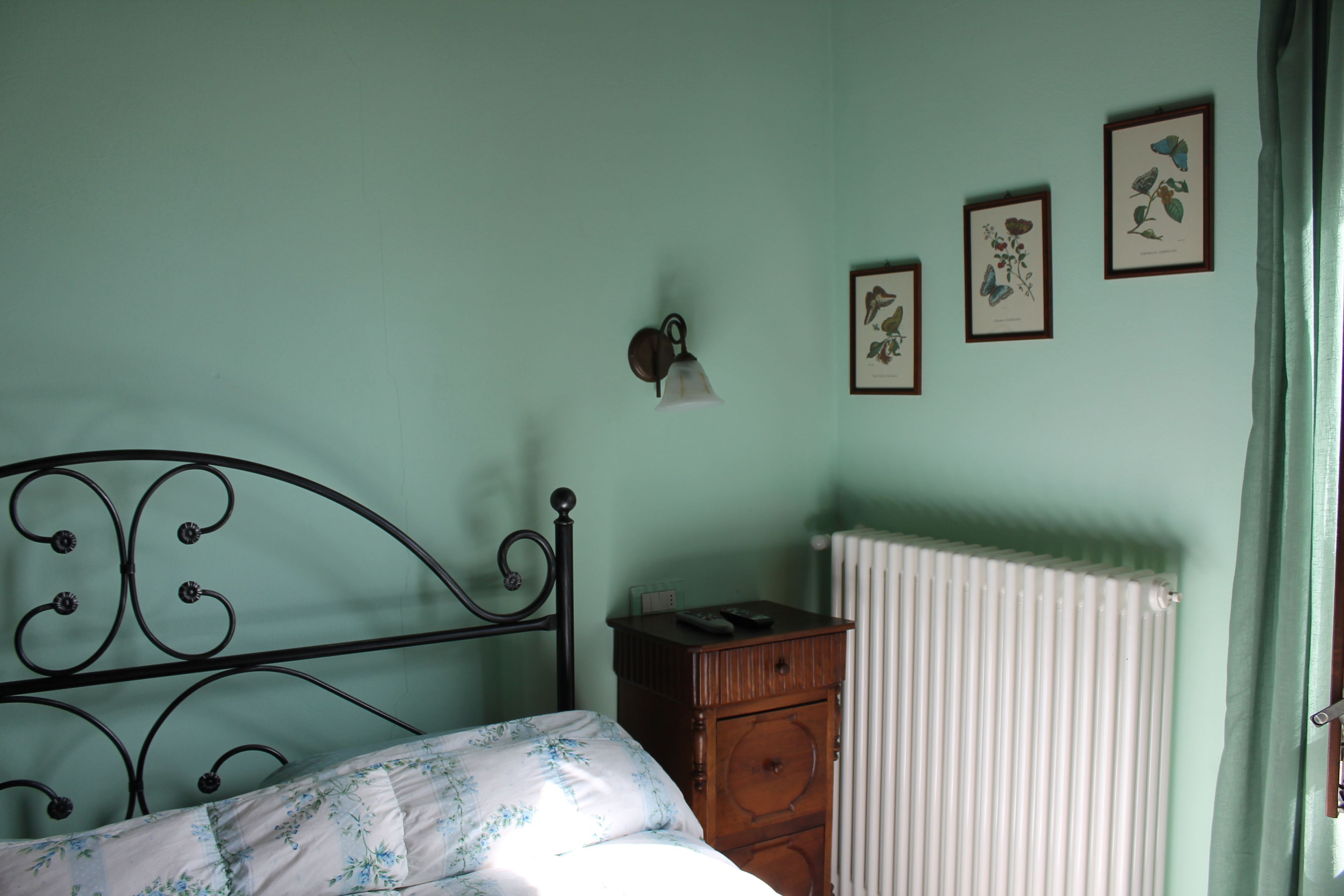 A bedroom with a metal bed frame, a wooden nightstand, a radiator, and framed botanical prints on the wall.