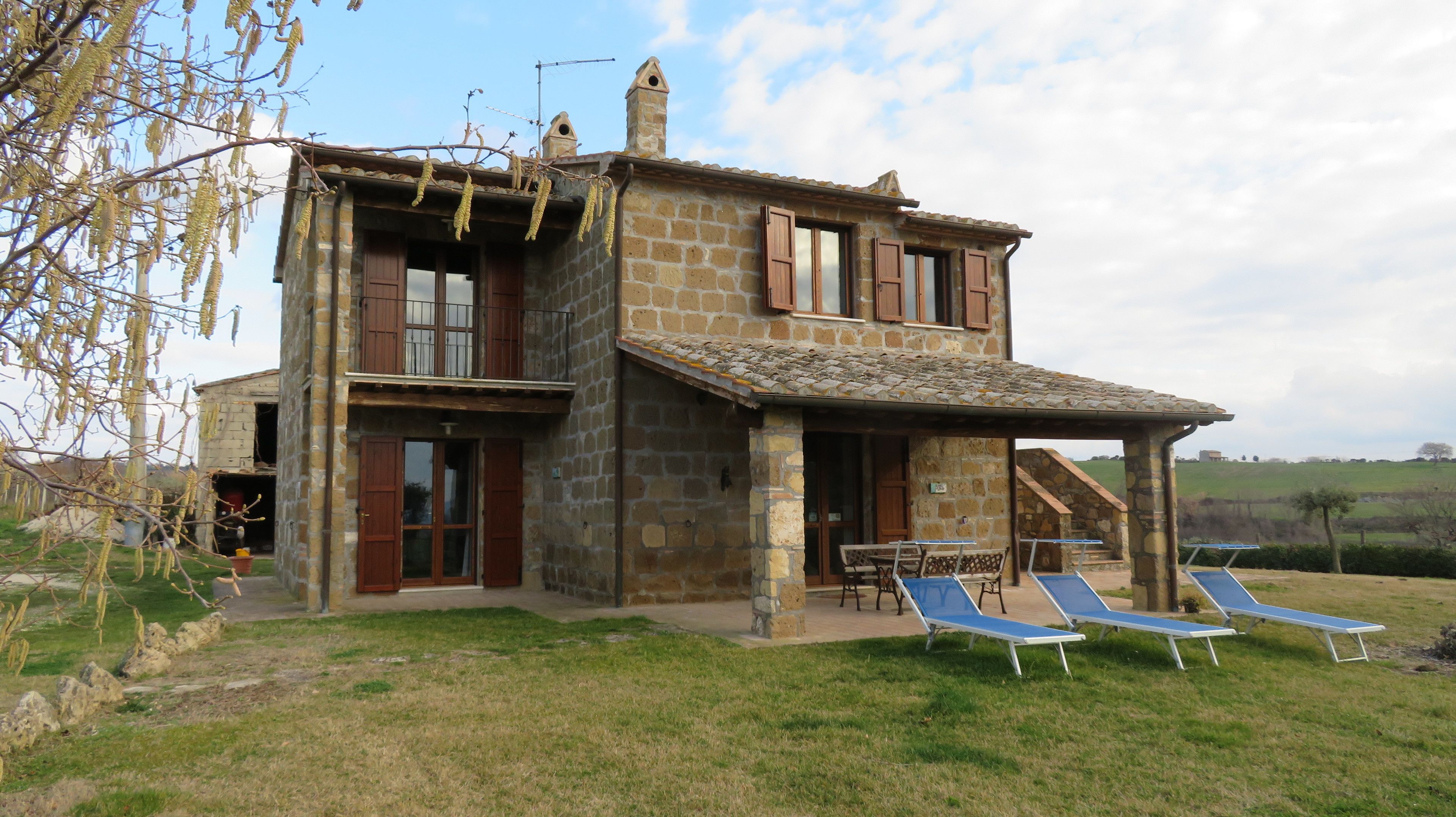 A stone house with a balcony, a small staircase, and two lounge chairs in the yard.