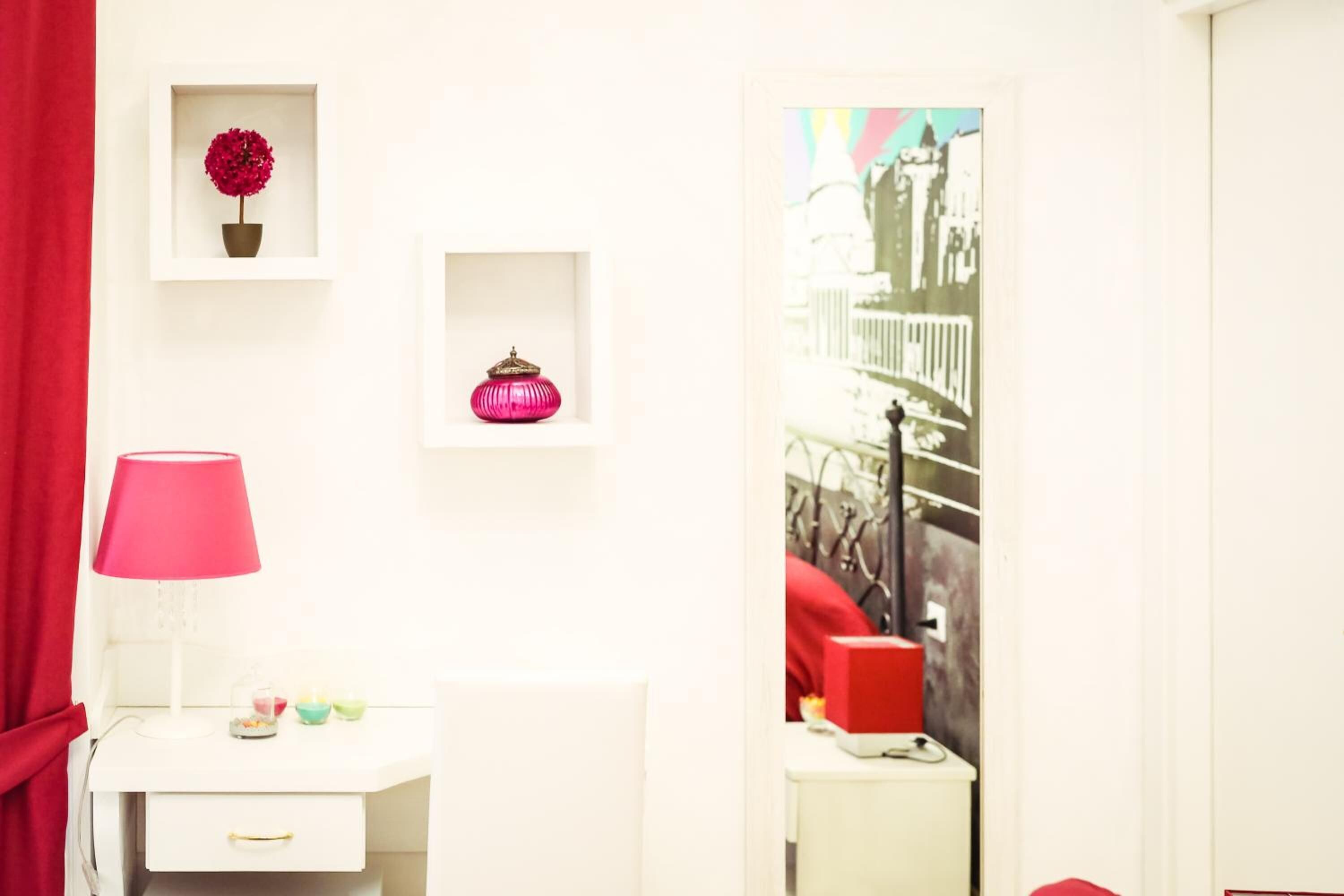 A white room with a red curtain, a white desk with a pink lamp, a small shelf with a red flower and a pink ornament, and a framed picture of a bridge.