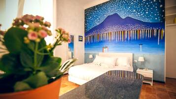 Une chambre d’hôtel avec un lit, des tables de chevet, une petite table, une plante en pot et un grand tableau mural.