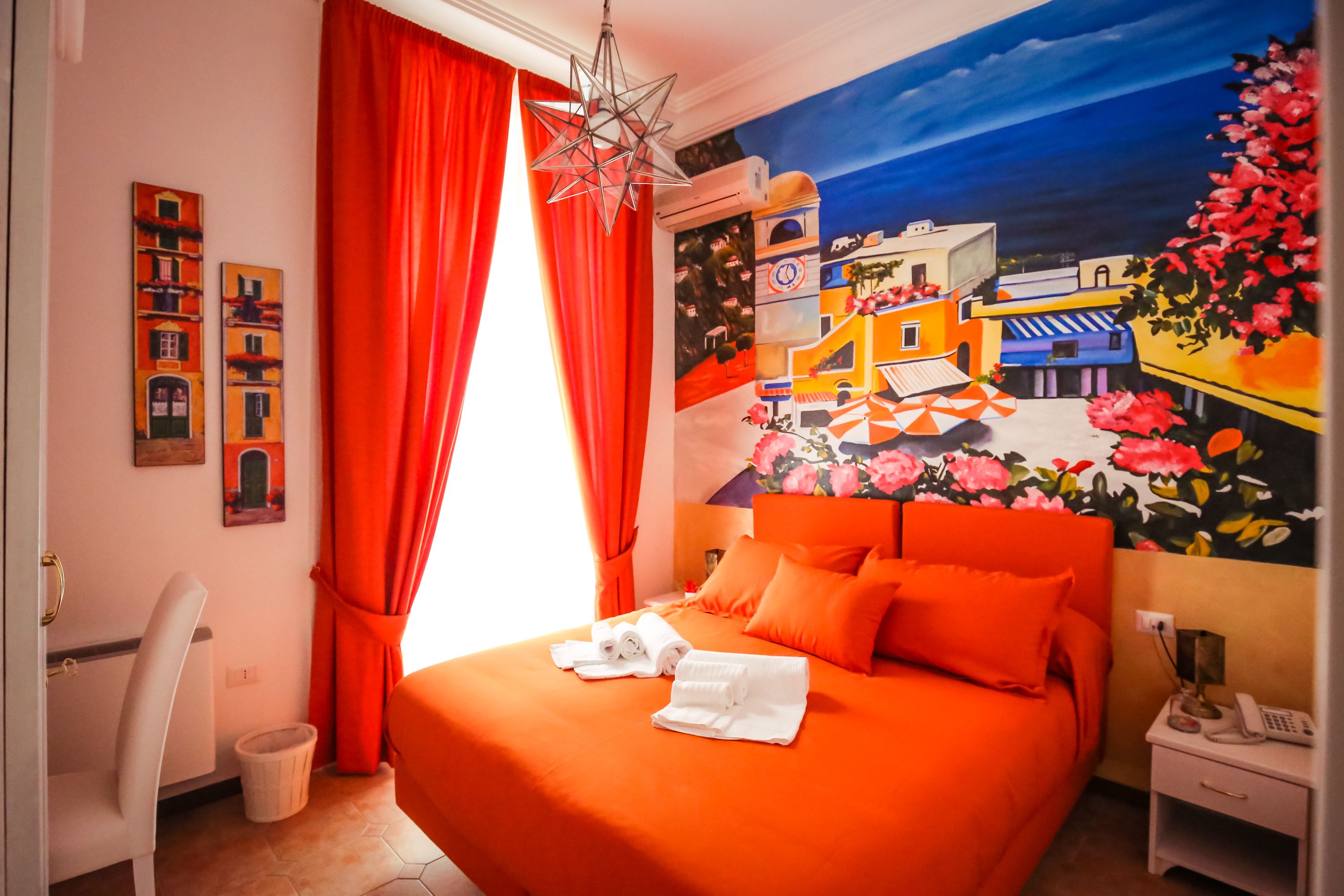 A hotel room with a large bed, orange bedding, a mural on the wall, and a red curtain.