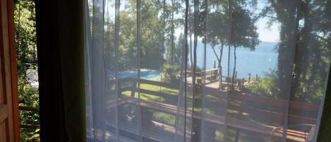 Vue depuis une chambre dotée d’une grande fenêtre donnant sur une piscine, une terrasse et des arbres.