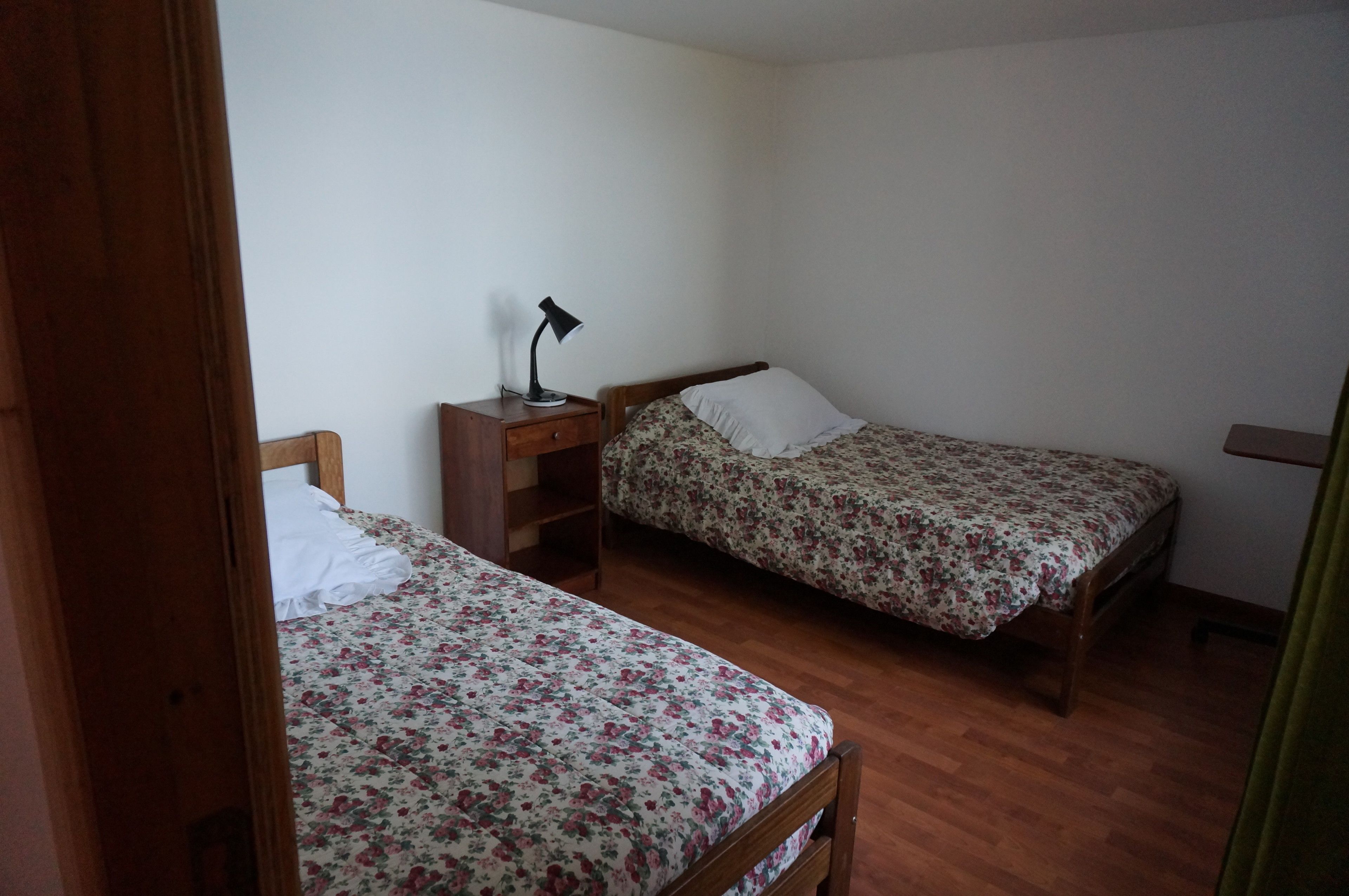 A bedroom with two single beds, wooden headboards, a nightstand with a lamp, and a wooden wardrobe.