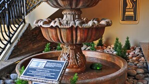 A multi-tiered fountain with a plaque, surrounded by rocks and small plants, in a stone-walled area with a staircase in the background. - Jockey Club Suites (Las Vegas)