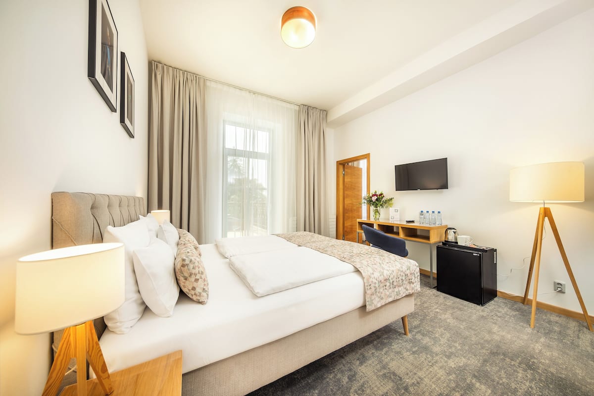 Een hotelkamer met een bed, nachtkastje, lamp, bureau, stoel en een televisie aan de muur.
