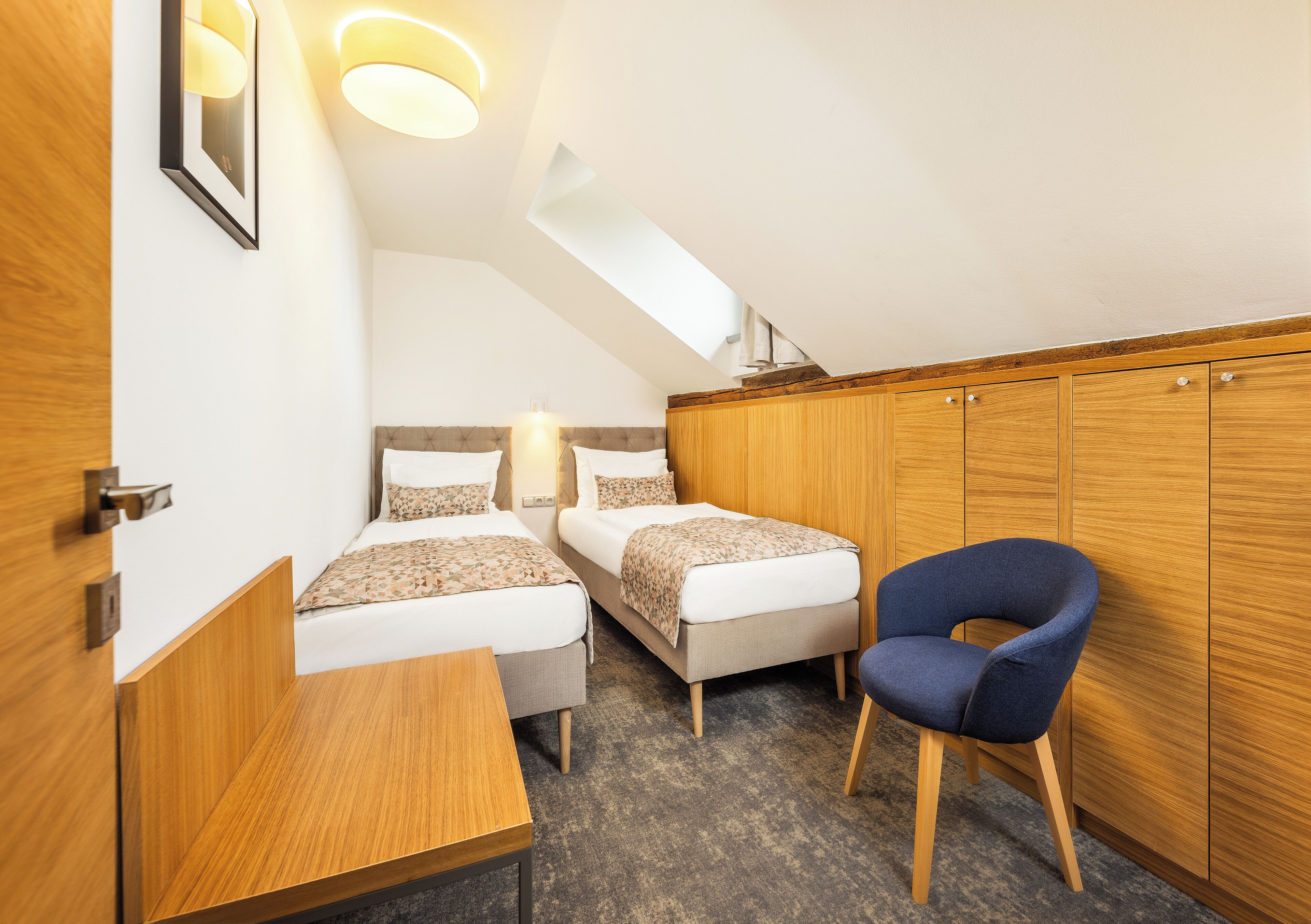 A compact room with two beds, a wooden wardrobe, a desk, and a blue chair.