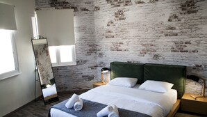 A modern bedroom with a brick wall, a large bed with white linens, two bedside tables, a mirror, and a window. - The Athens Version Luxury Suites (Athens)