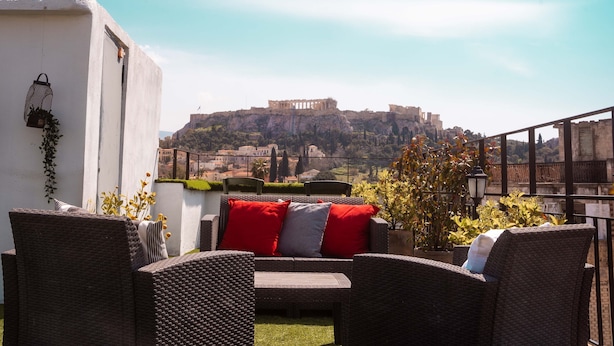 An outdoor seating area with wicker furniture and red cushions, overlooking a historic building on a hill. - The Athens Version Luxury Suites (Athens)