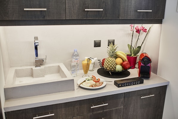 A kitchen with a white countertop, a sink, and a fruit bowl containing bananas, pineapples, and apples. There is a water bottle, a glass of juice, and a red appliance on the counter. - The Athens Version Luxury Suites (Athens)