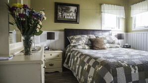 Un dormitorio con cama, mesitas de noche, un espejo, un cuadro enmarcado y un florero con flores.
