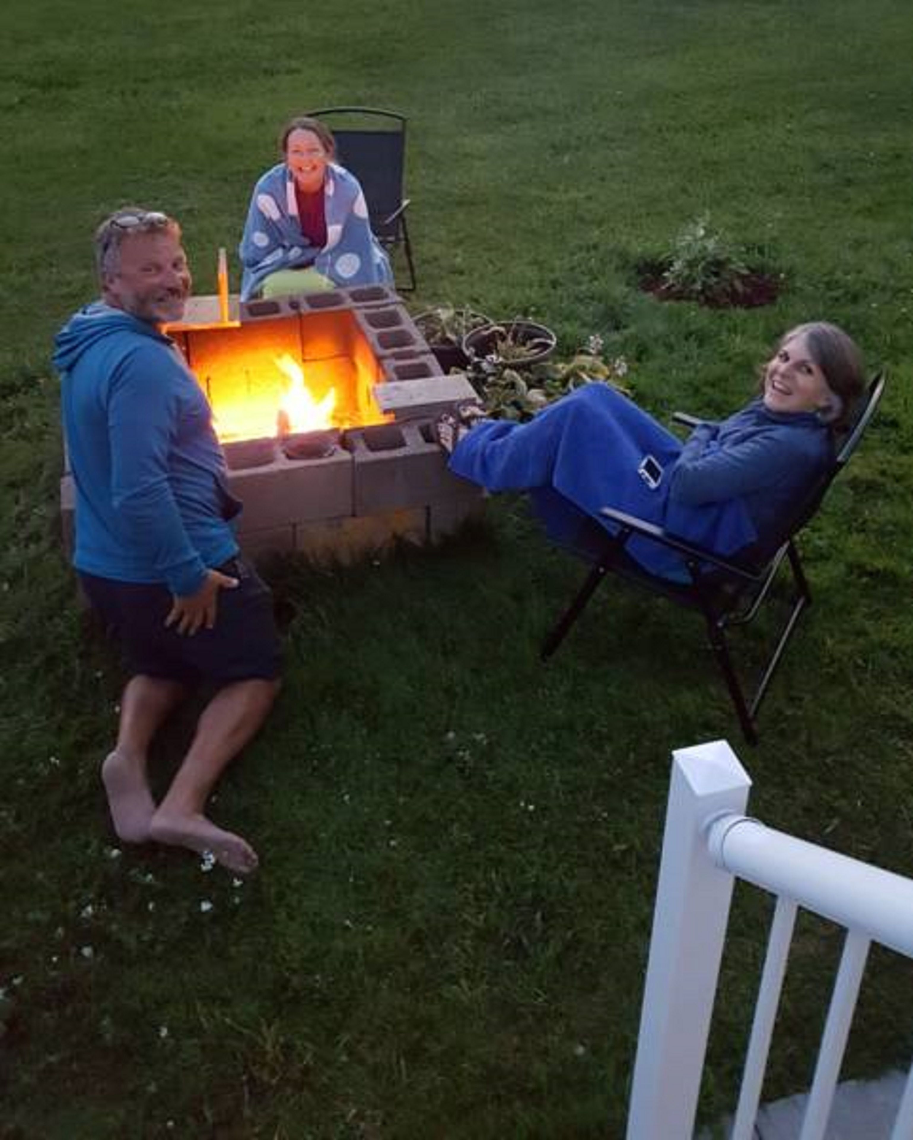 Three people sitting around a fire pit in an outdoor setting with a grassy lawn and a white railing.