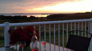 Una mesa con una botella y dos vasos, un jarrón con flores rojas, y vista al mar al atardecer.