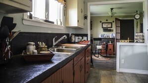Una cocina con armarios de madera, una encimera negra y una ventana con cortina escocesa. Un letrero sobre la puerta dice: “Bless All Who Enter Here”.