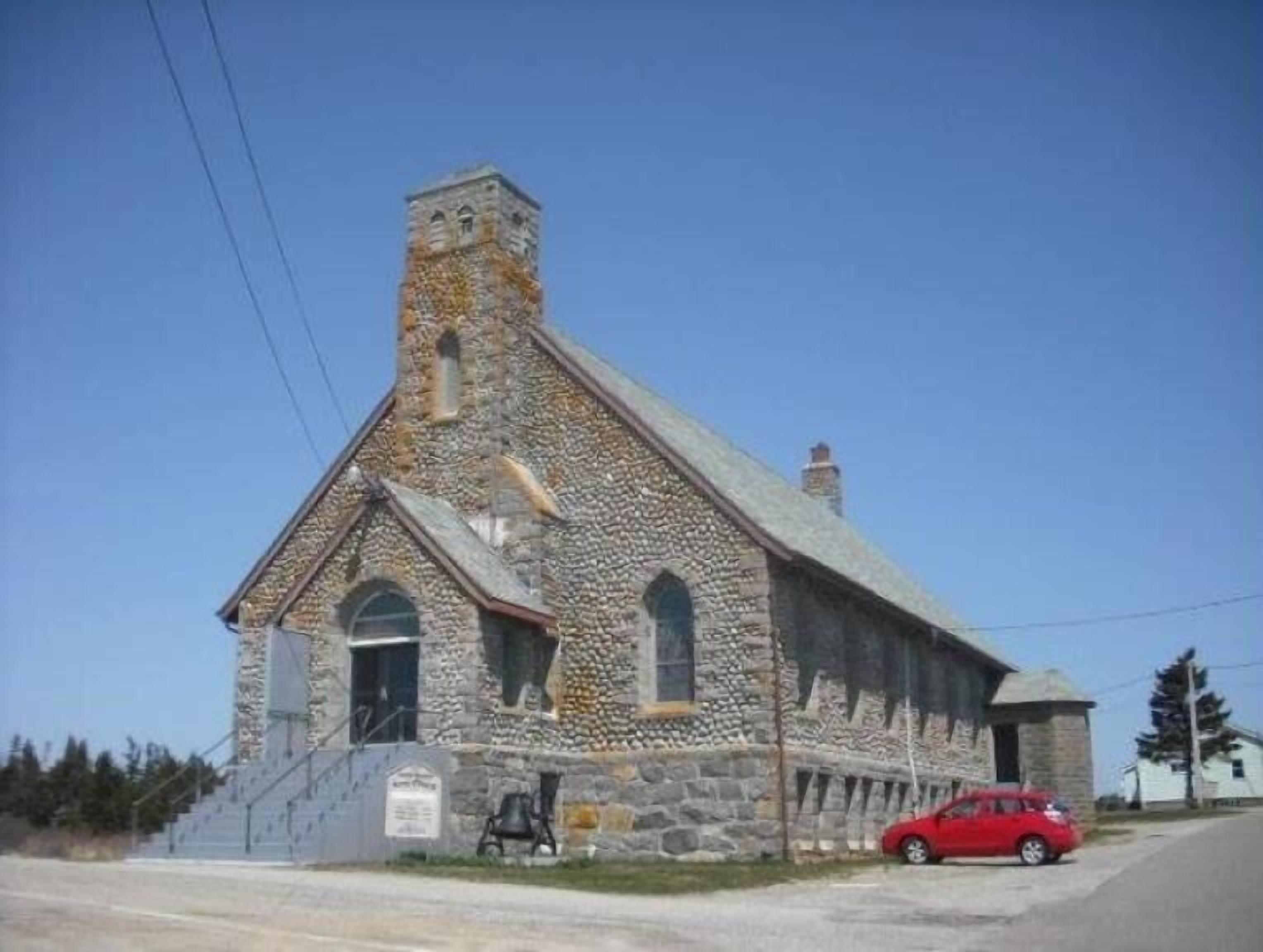 A stone church with a bell tower, a small porch, and a red car parked in front.