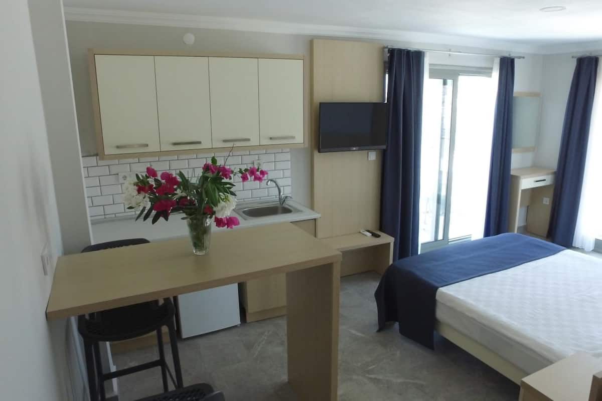 A compact hotel room with a kitchenette, a bed, a TV, and a window with curtains.