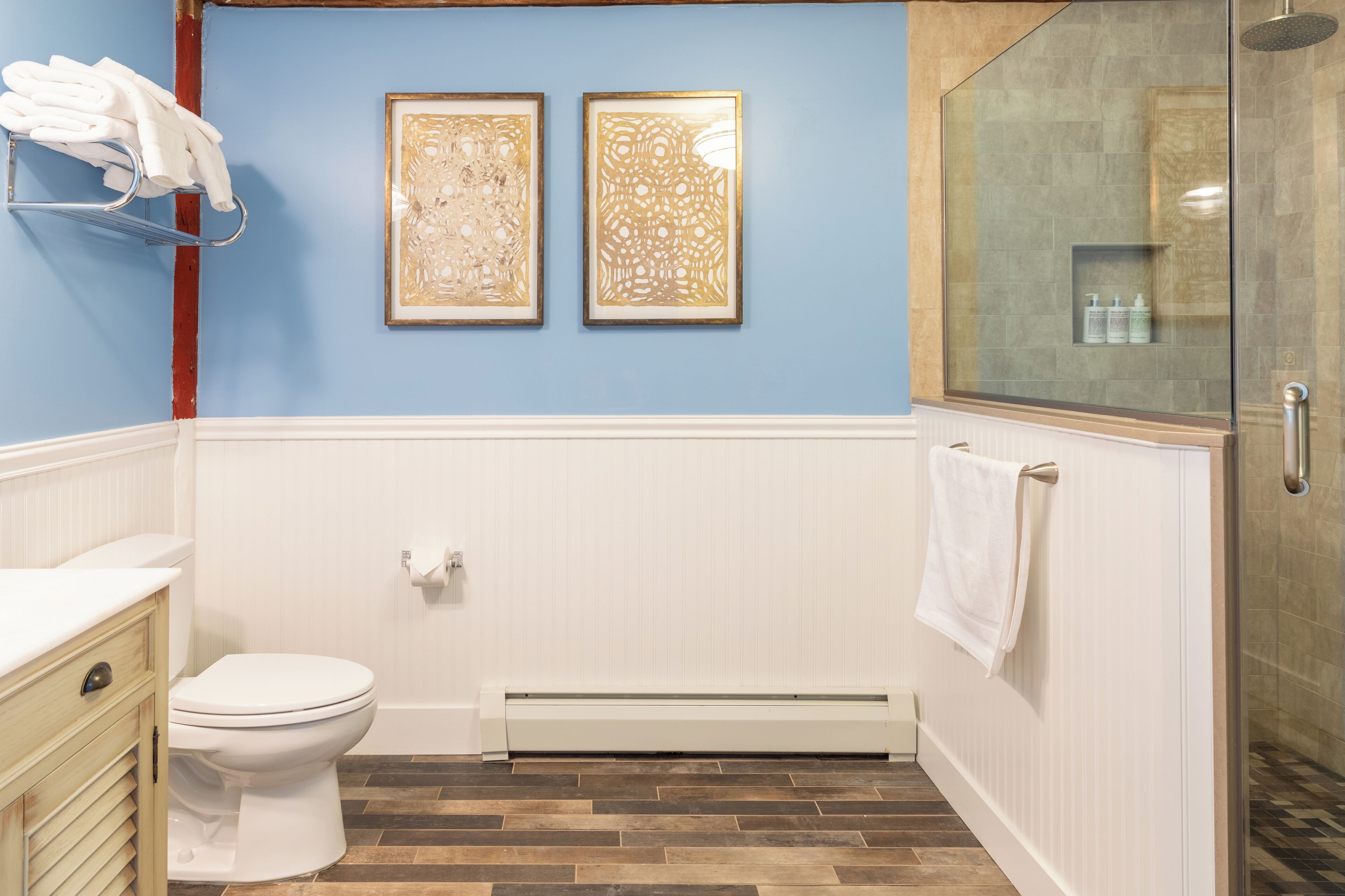 A bathroom with a white toilet, a towel rack with towels, two framed artworks on the wall, a glass shower enclosure, and a radiator.