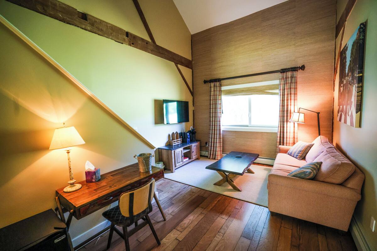 A cozy room with a wooden desk, a cushioned sofa, a flat-screen TV, and a window with curtains.