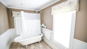 Une salle de bain avec une baignoire sur pieds, un rideau de douche, une fenêtre avec des stores et un radiateur.