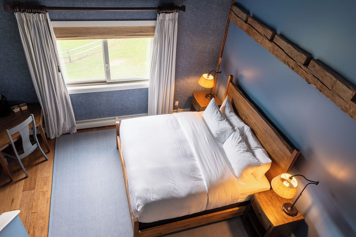 A neatly made bed with white linens, a wooden headboard, and two bedside lamps. A window with curtains is visible, and a small table with a chair is in the corner.