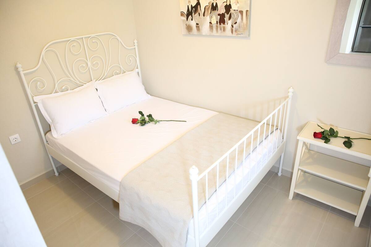 A white metal bed with a decorative headboard, a white bedside table with a rose, and a wall-mounted artwork.