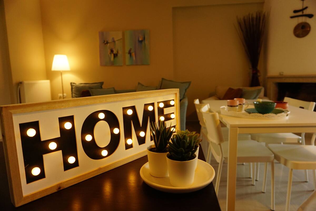 A room with a 'HOME' sign, a dining table, chairs, a lamp, and two potted plants.