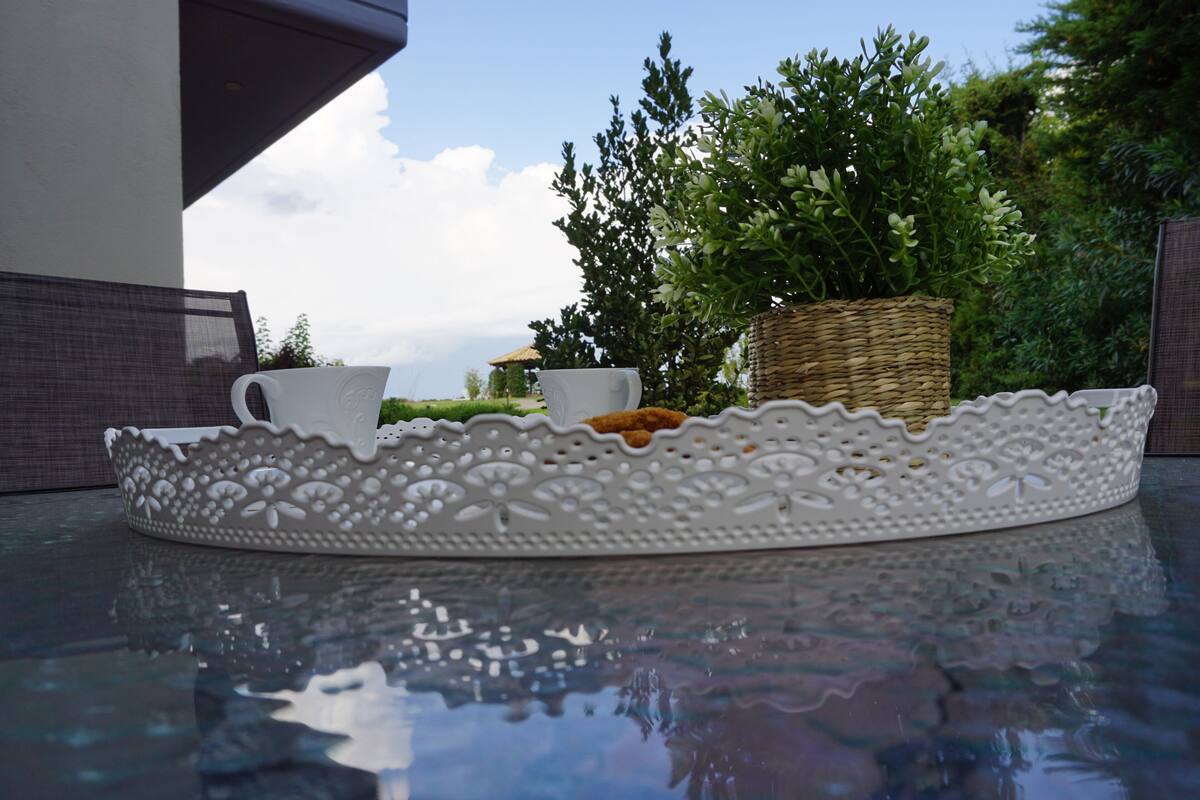 A white decorative tray with a woven basket on a reflective surface, surrounded by greenery and a building in the background.