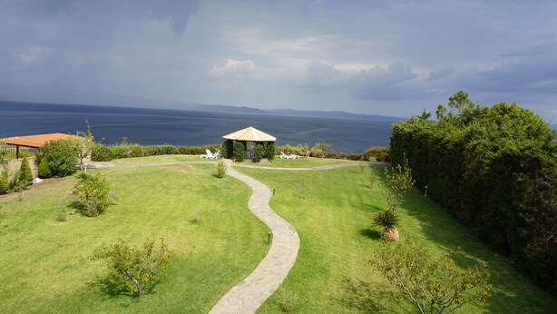 Ein grünes Areal mit einem Kiesweg, der zu einem Gazebo führt, umgeben von Bäumen und mit Blick auf ein Gewässer.