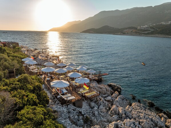 A coastal restaurant with outdoor seating overlooking the sea, wooden deck, and umbrellas. - Suna Sun Hotel (Kas)