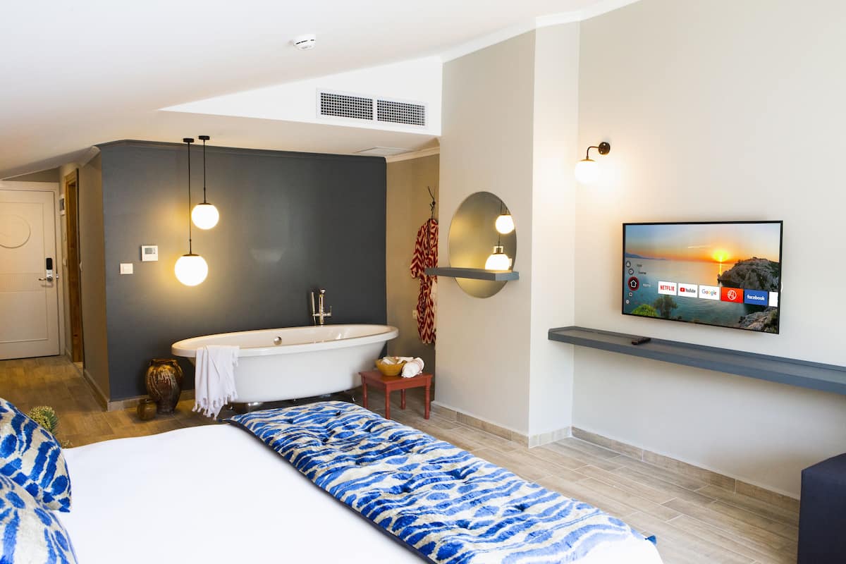 A modern hotel room with a large bed, a bathtub, a television mounted on the wall, and a small table with a vase.