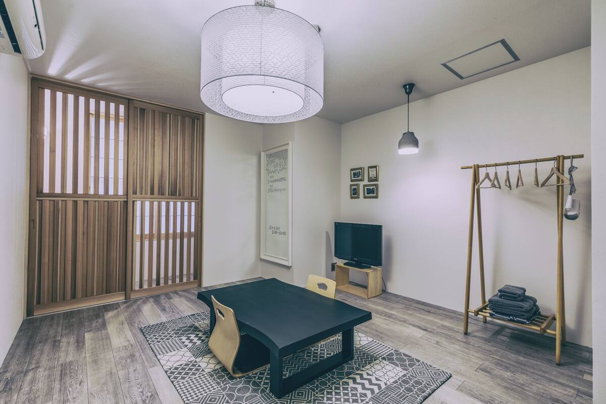 Una habitación con una mesa negra, un televisor sobre un soporte, una lámpara colgante, una partición de madera y un tendedero.