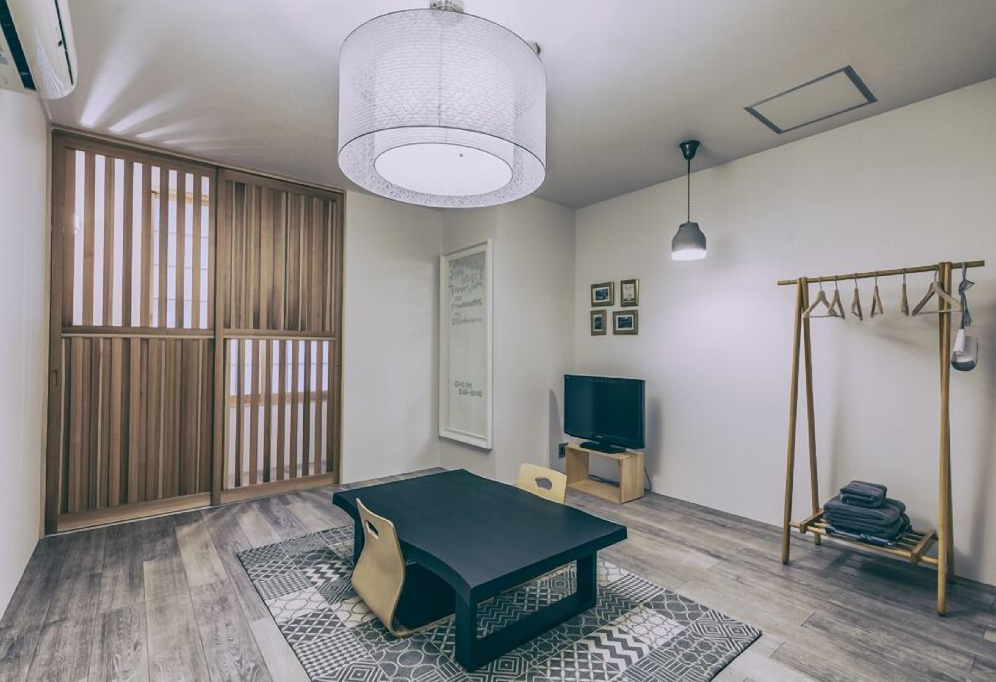 Una habitación con una mesa negra, un televisor, una lámpara colgante, un separador de madera y un perchero.