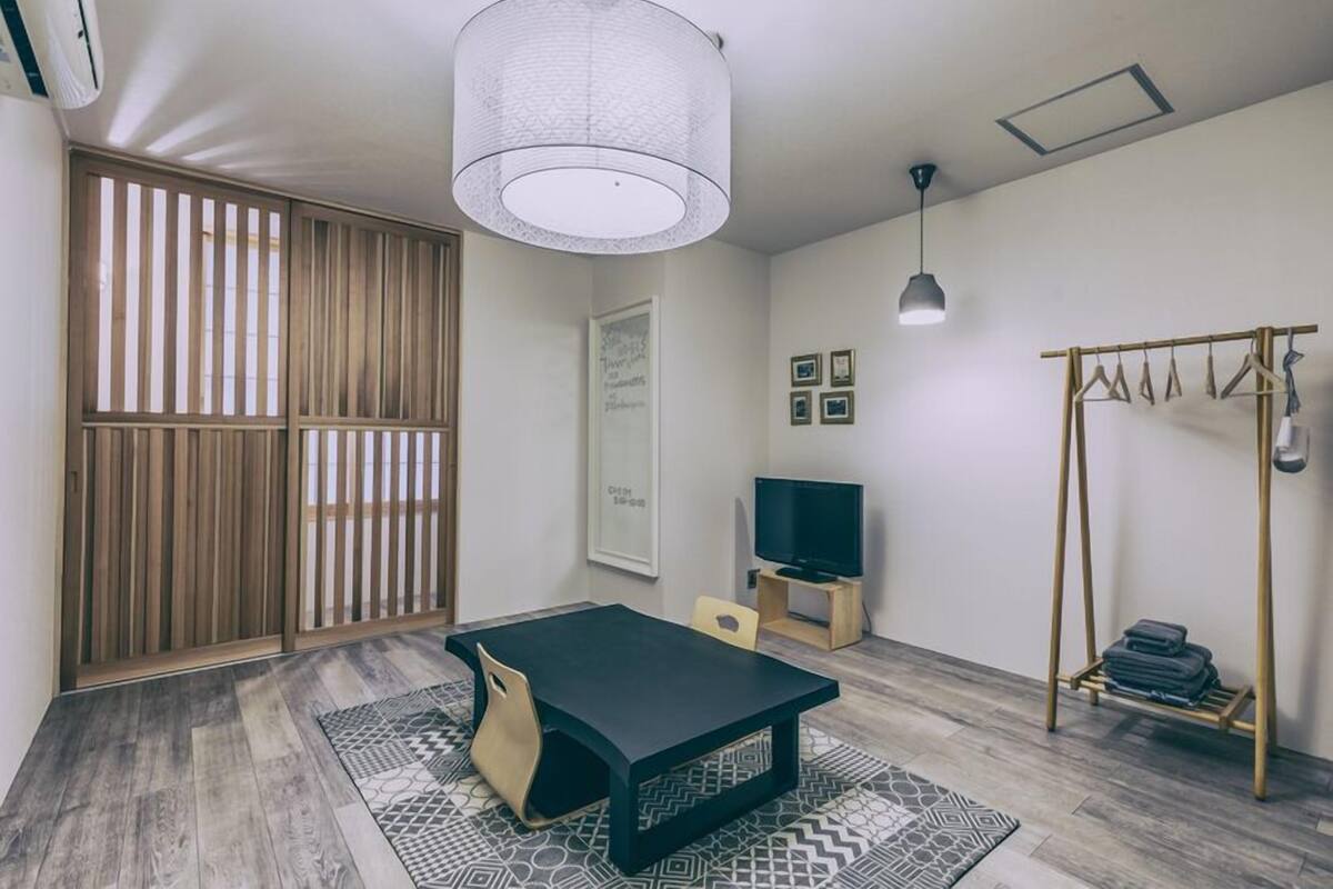 Una habitación con una mesa negra, un televisor, una lámpara colgante, un separador de madera y un perchero.
