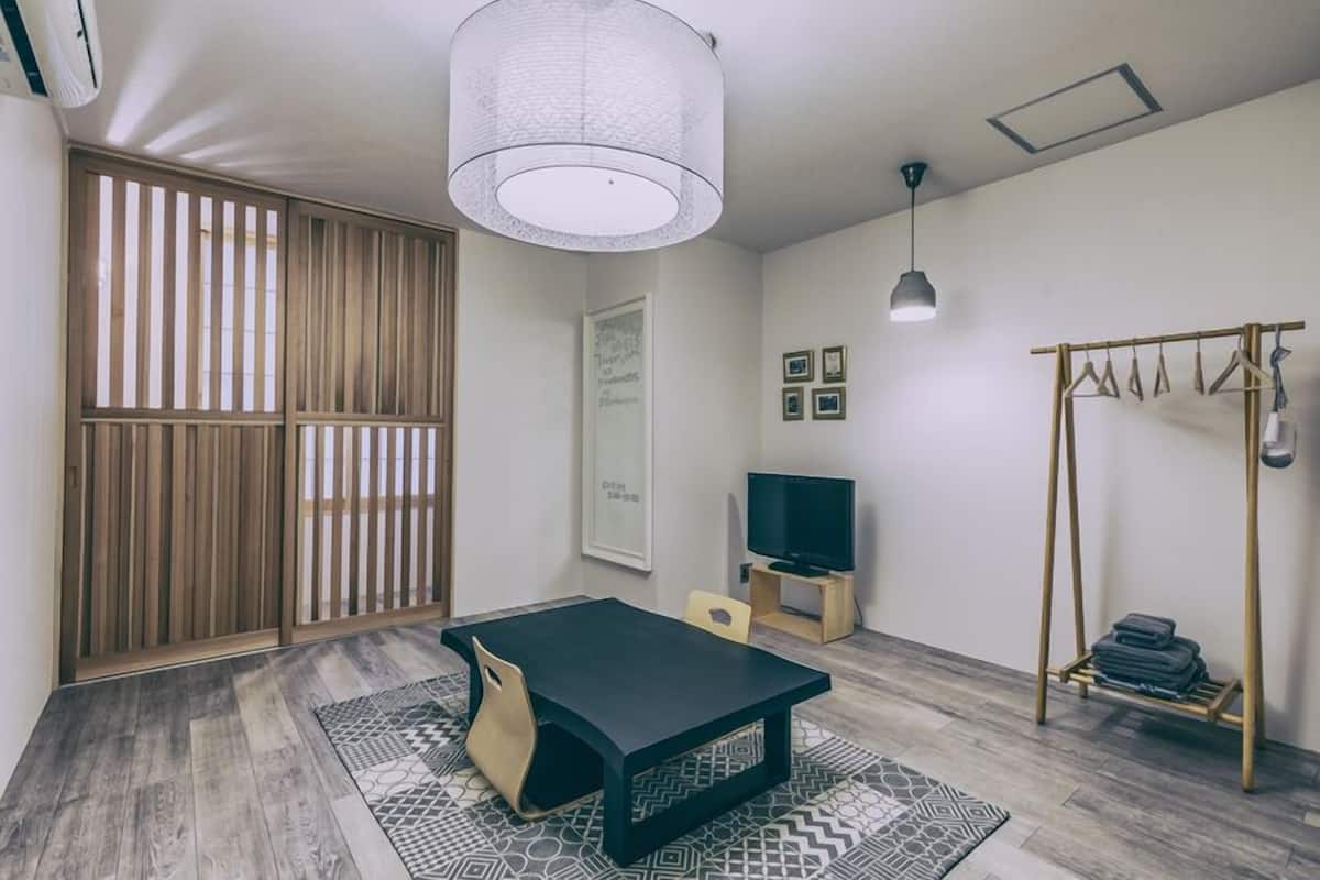 Una habitación con una mesa negra, un televisor, una lámpara colgante, un separador de madera y un perchero.