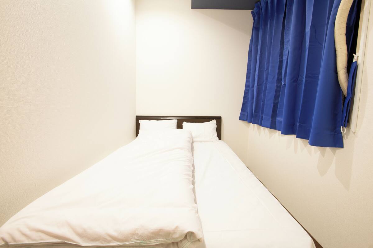 A single bed with white bedding in a room with a plain wall and a blue curtain on the right.