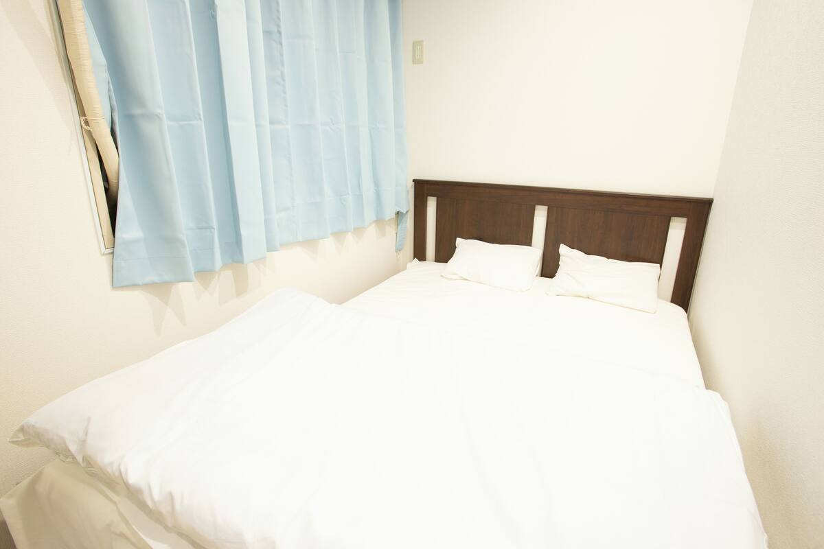 A neatly made bed with white linens in a simple room with a wooden headboard and light blue curtains.