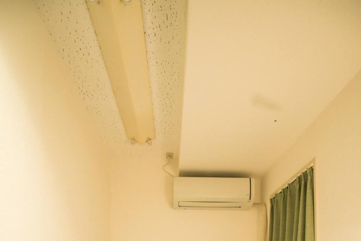 A small, single-bed room with a plain white wall, a window with green curtains, and an air conditioning unit mounted on the wall.