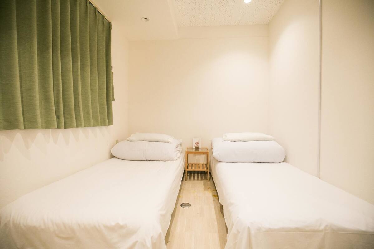 A small, clean room with two single beds, a wooden nightstand, and a green curtain.