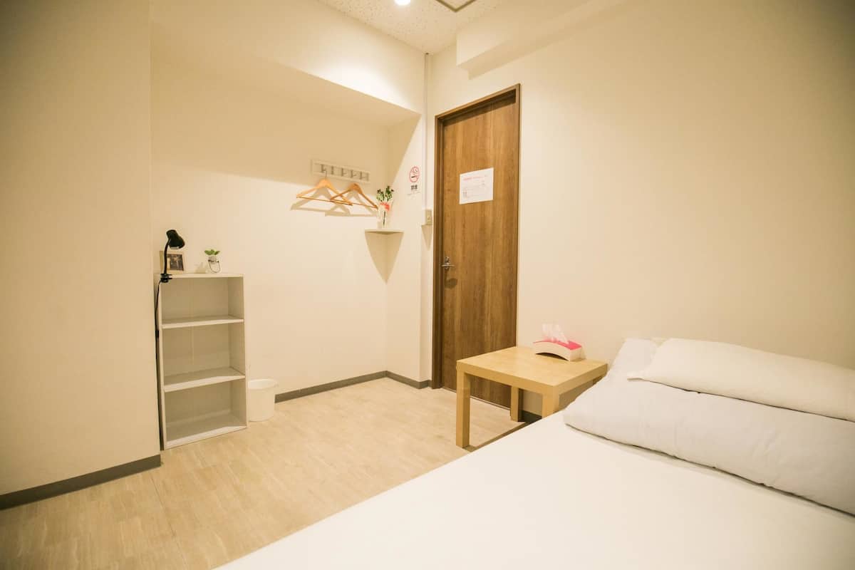 A bedroom with a bed, a small table, a wooden door, and a shelf with a hanging plant.