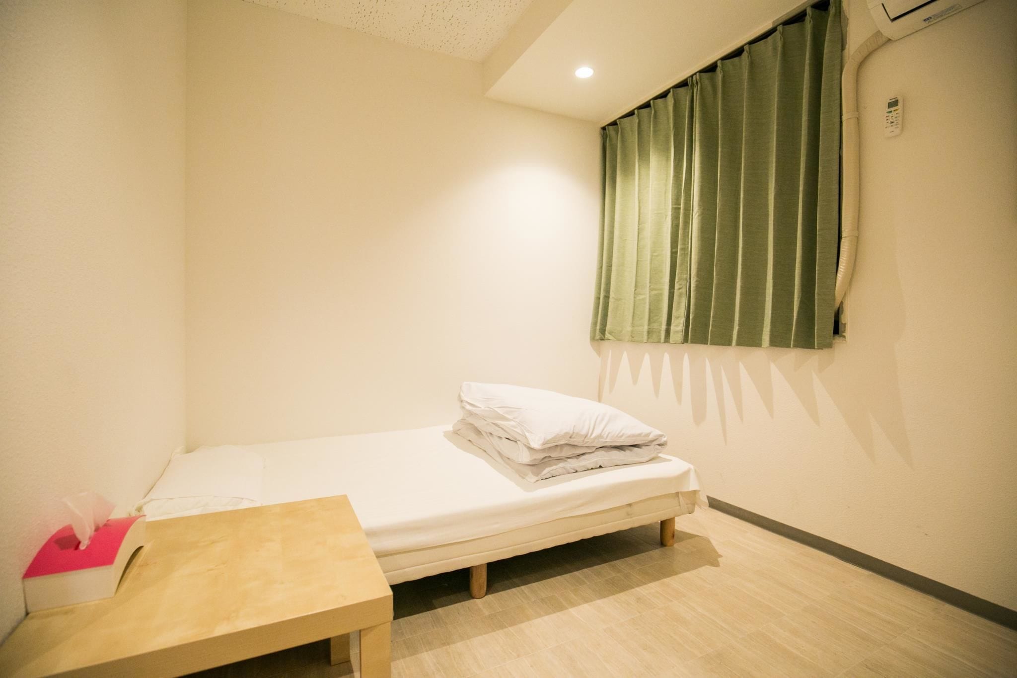 A small, single-bed room with a wooden table, a green curtain, and a wall-mounted air conditioning unit.