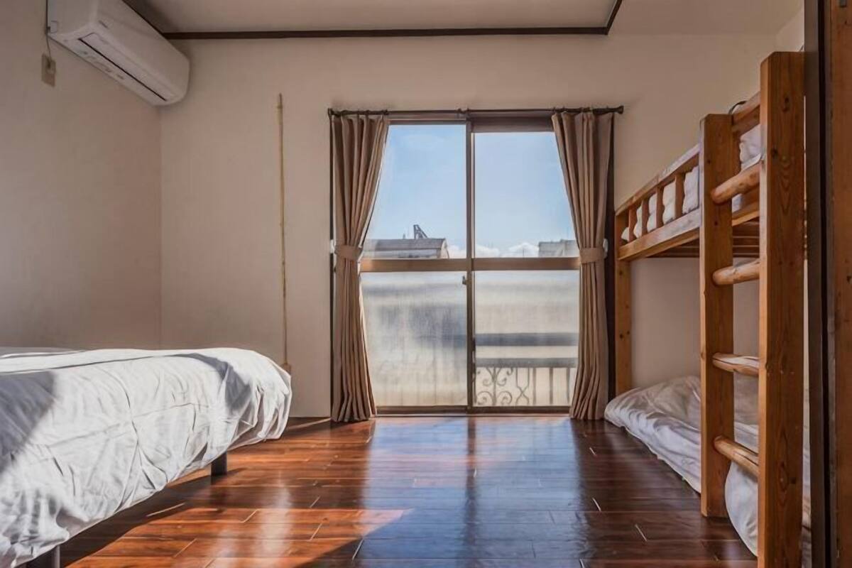 A room with a bunk bed, a single bed, a large window with curtains, and a view of the city.