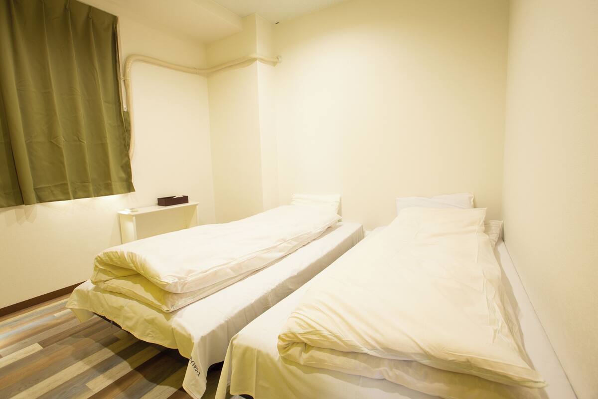 A small, clean room with two single beds, a bedside table, and a window with a green curtain.