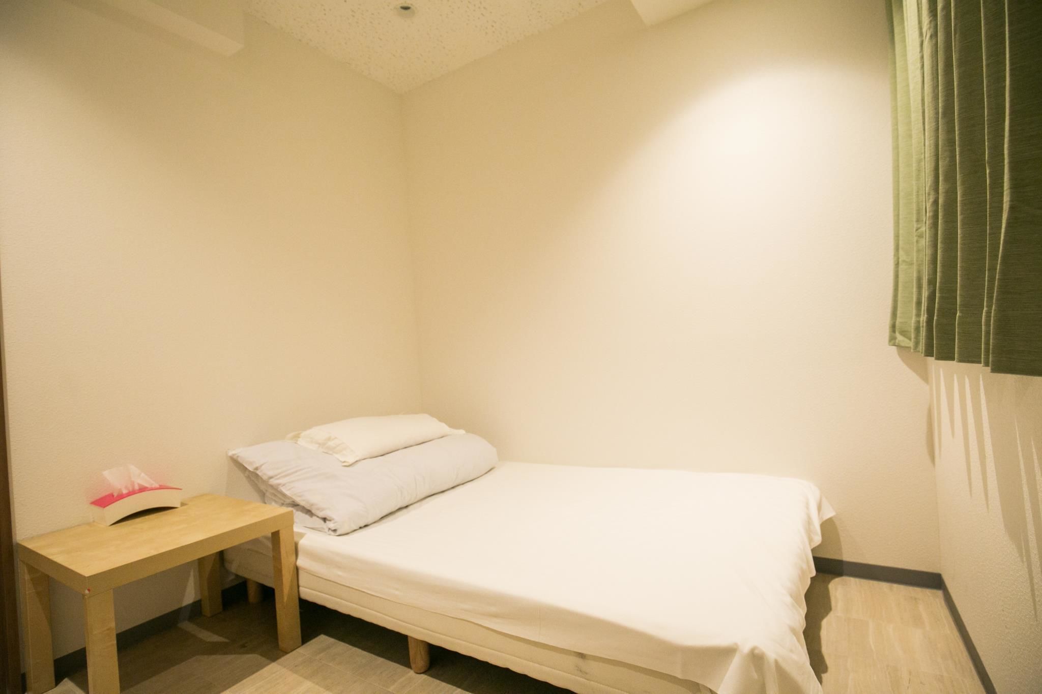 A small, single-bed room with a wooden nightstand, a white bedspread, and a green curtain on the window.