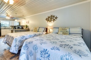 A bedroom with two beds, a ceiling fan, a kitchen in the background, and decorative pillows. - 1 Mi to Pier: Ocean Isle Beach Studio w/ Patio! (Ocean Isle Beach)