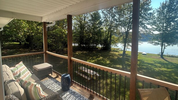 A covered deck with outdoor furniture, a view of a lake, and surrounding greenery.