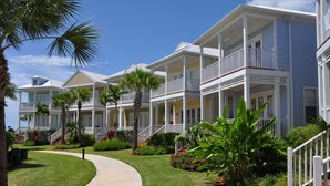 Een rij strandhotels in kuststijl met meerdere balkons, een verhard voetpad en weelderige beplanting.