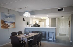 A dining area with a wooden table and chairs, a kitchen with white cabinets, and a pendant light. - 306 SEASCAPE CONDOMINIUMS BEACH SIDE CONDO (South Padre Island)
