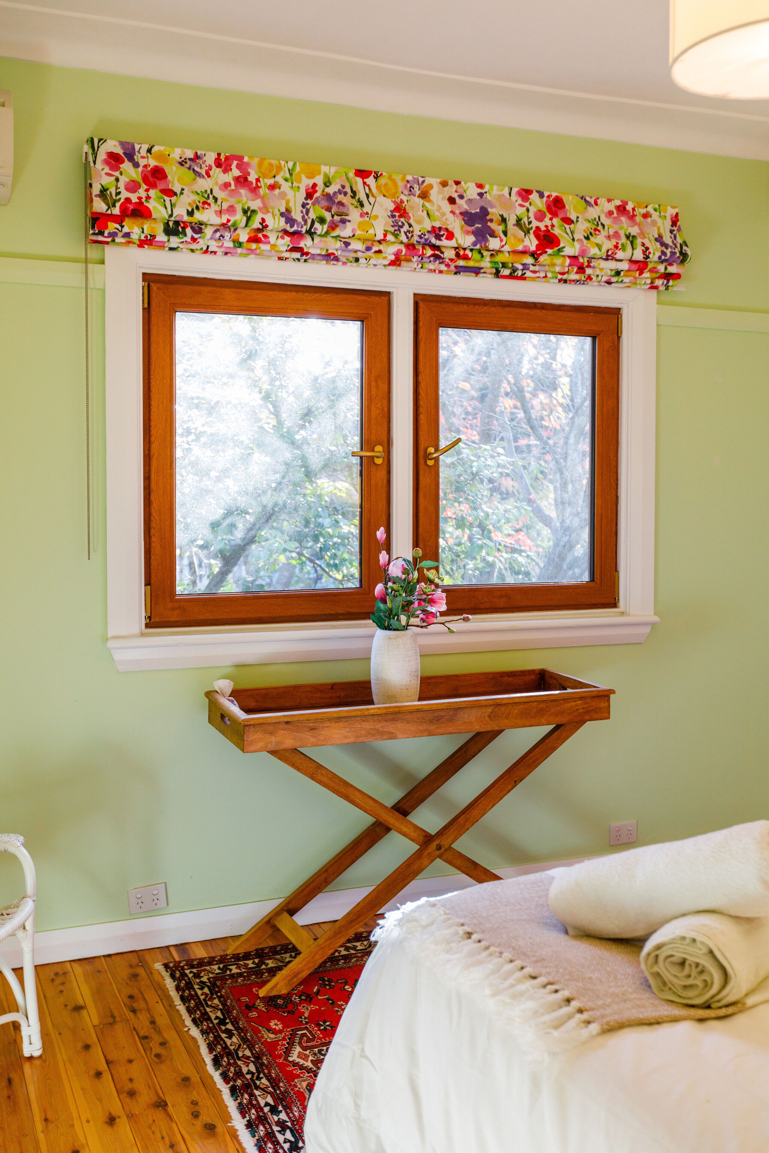 A room with a bed, a window with a floral valance, a wooden side table with a vase of flowers, and a red patterned rug.