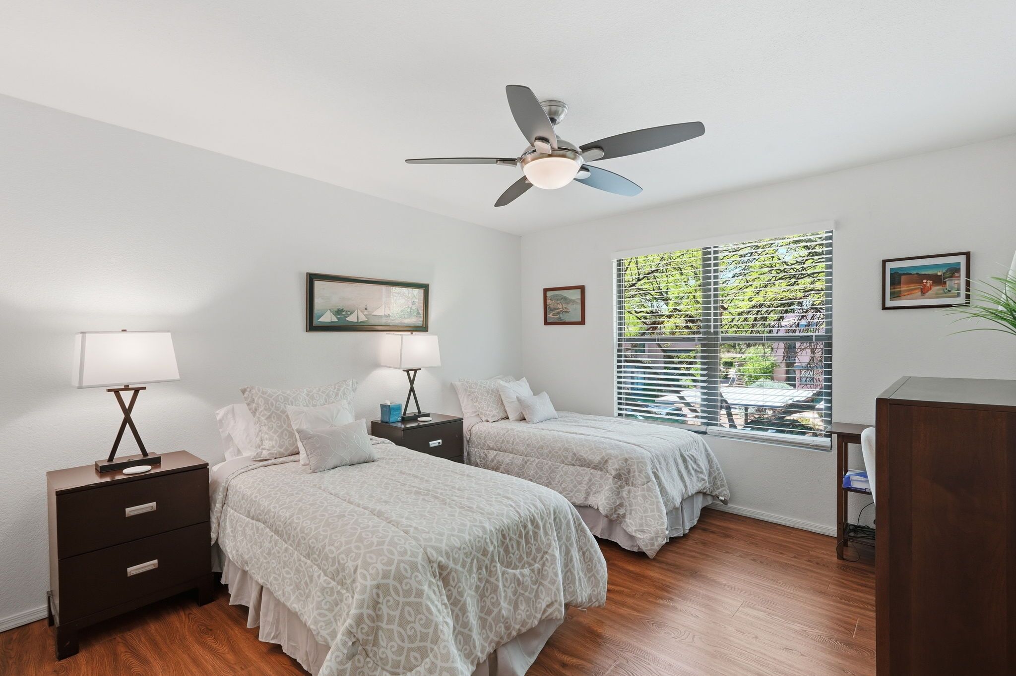 Une chambre avec deux lits, un ventilateur de plafond, une table de chevet avec une lampe, une fenêtre avec des stores et un bureau avec une chaise.