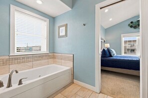A bathroom with a bathtub, a window with blinds, and a door leading to a bedroom with a bed.
