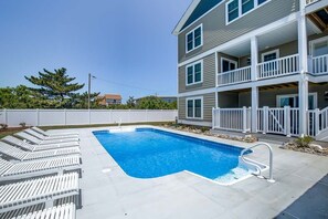 A rectangular swimming pool with a white deck and lounge chairs, surrounded by a white fence and a multi-story building with balconies.